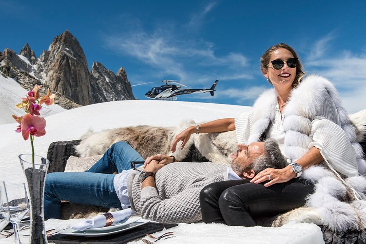 A man and woman are laying in the snow with a helicopter in the background