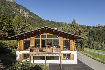 A beautiful wooden chalet with a balcony, set against a backdrop of forested mountains and a clear blue sky.