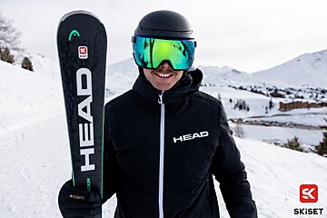 A man wearing a head jacket holds a pair of skis
