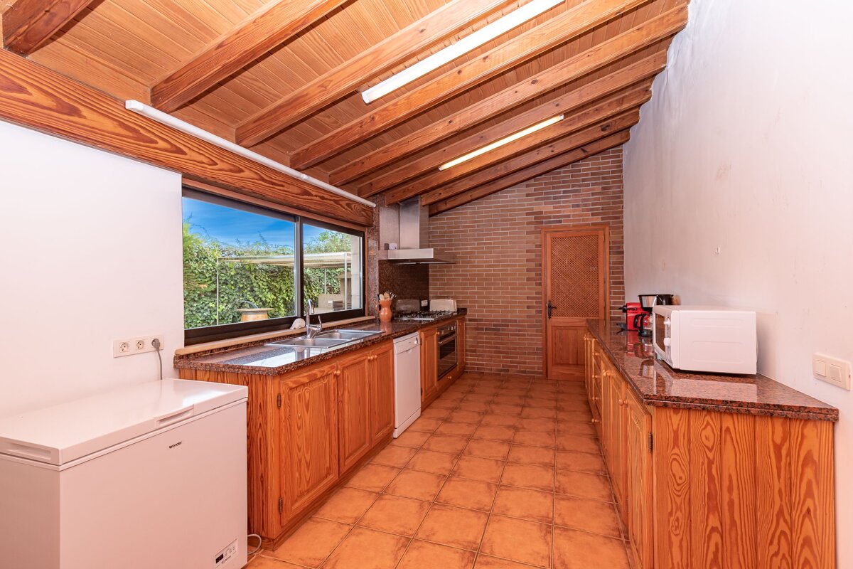 Rustic kitchen with a high wooden beamed ceiling, brick accent wall, and terracotta tiled floor. Wooden cabinets line the space with a garden view.