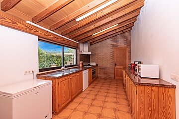 Rustic kitchen with a high wooden beamed ceiling, brick accent wall, and terracotta tiled floor. Wooden cabinets line the space with a garden view.