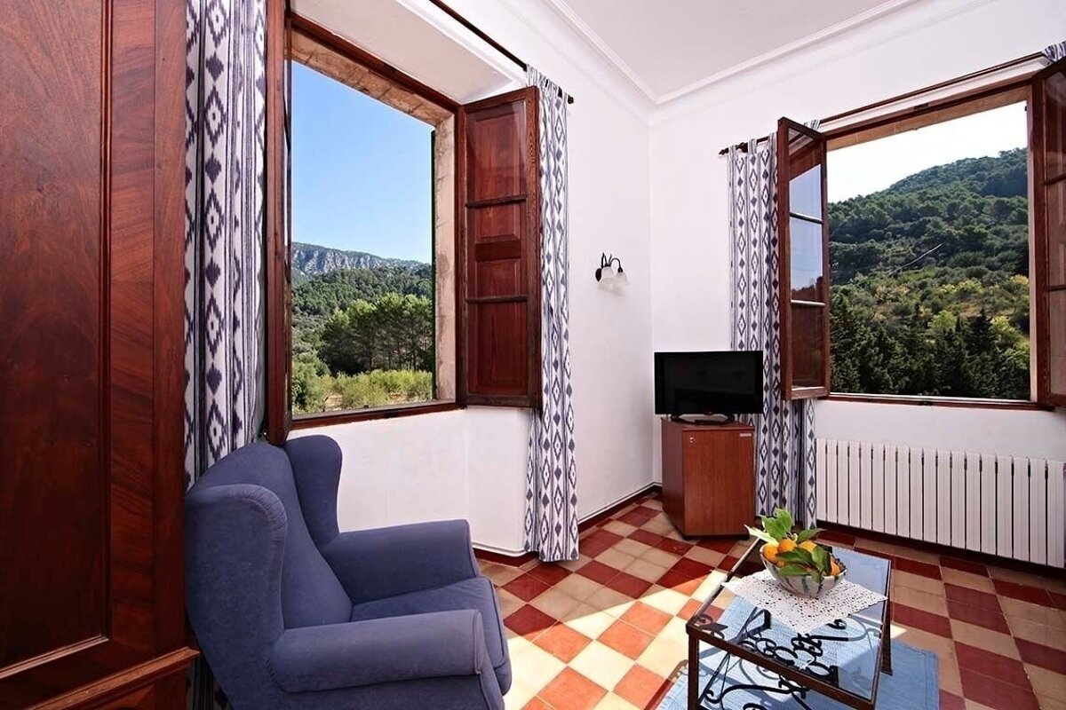 A rustic room features two windows with stunning mountain and forest views. It has a blue armchair, checkerboard tiled floor, and a small TV.