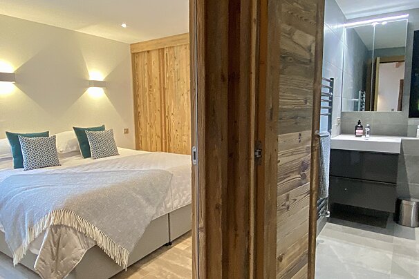 A cozy bedroom with a large bed and patterned pillows, and a modern en-suite bathroom with a vanity and mirror, separated by a rustic wooden door frame.
