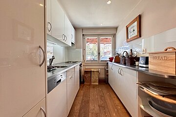 A bright, narrow kitchen with sleek white cabinets, a wooden floor, and a window overlooking snowy scenery. A sink, stove, toaster, and 