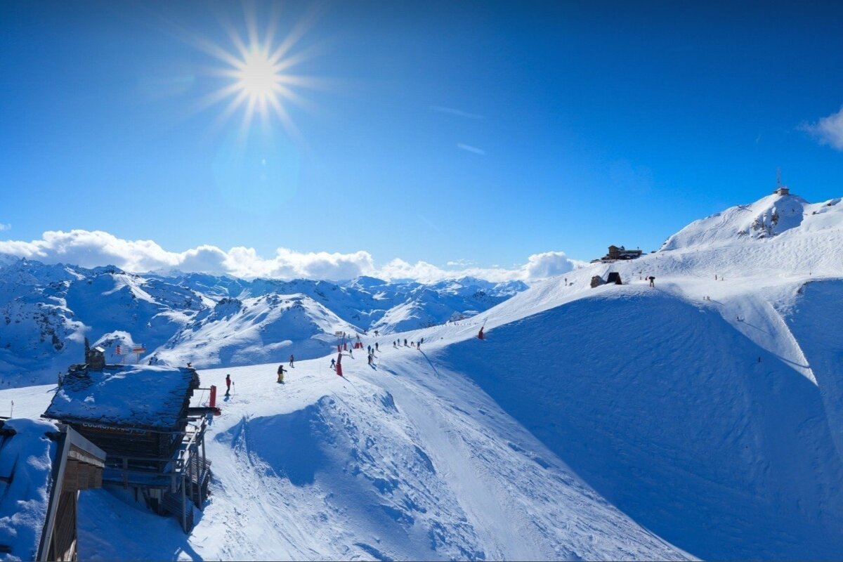 A bright, sunny day illuminates a vast snow-covered mountain ski resort. Skiers traverse the slopes, with buildings nestled among the peaks under a clear blue sky.