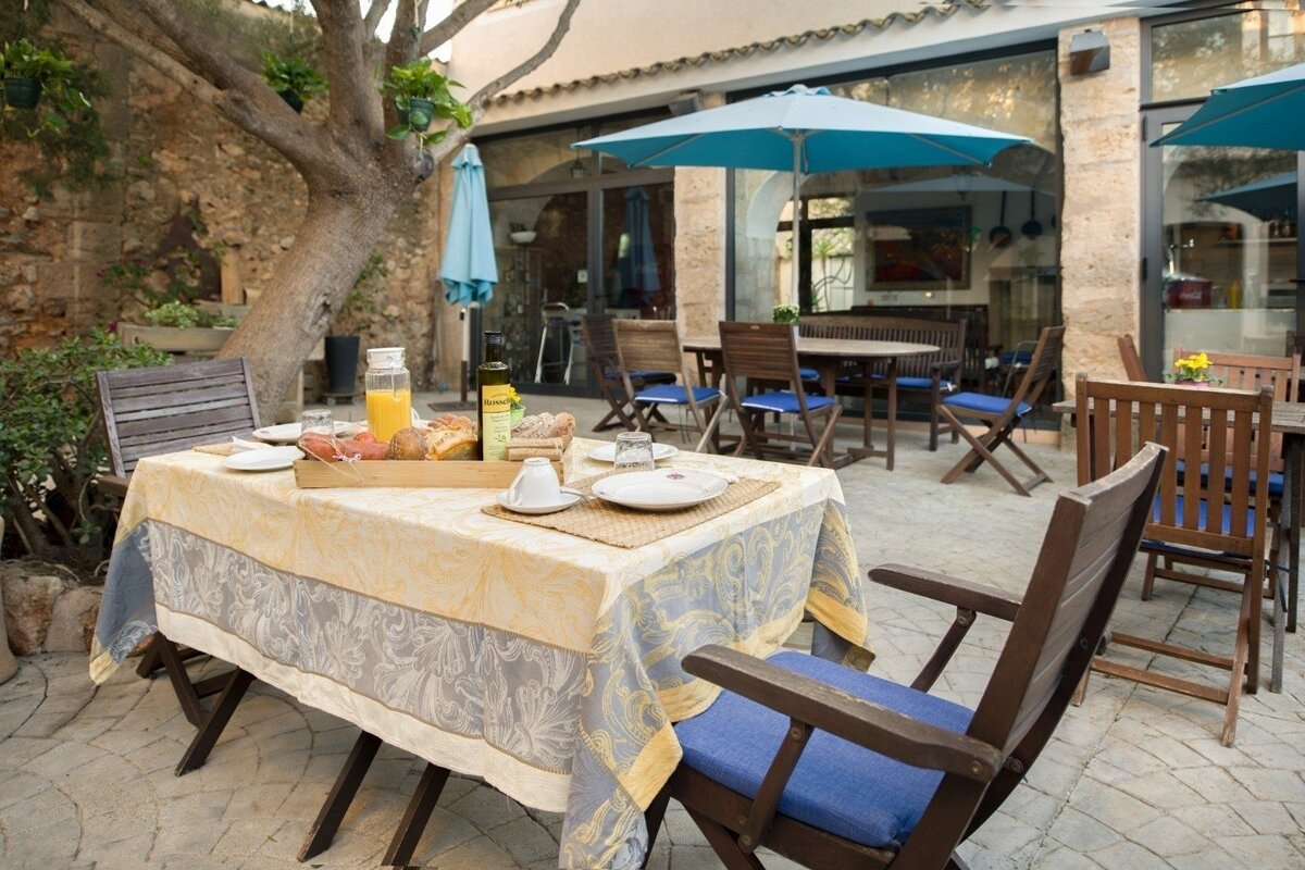 An outdoor cafe patio with a table set for breakfast, surrounded by trees and blue umbrellas, creating a relaxed atmosphere.