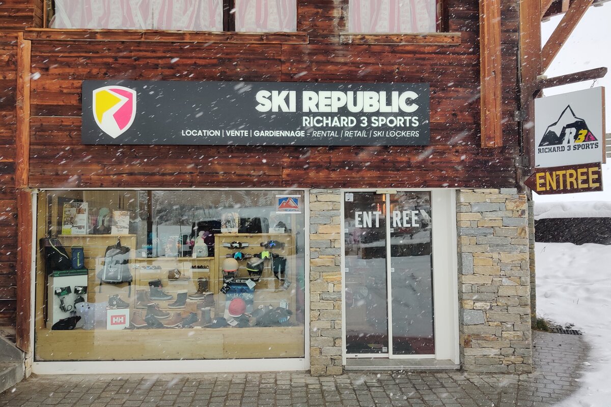 Snow falls on a ski shop with a wooden and stone facade. The sign reads 