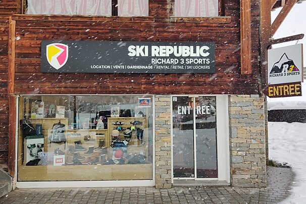 Snow falls on a ski shop with a wooden and stone facade. The sign reads 
