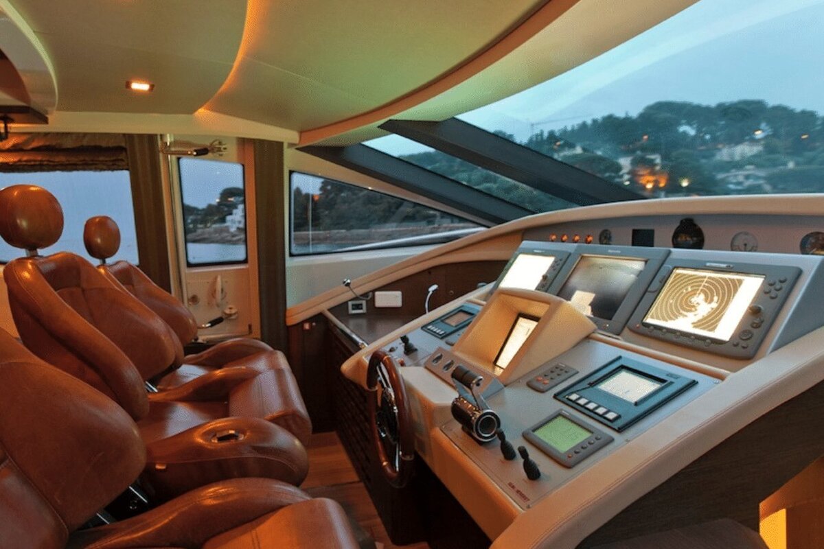 Plush yacht cockpit featuring brown leather chairs, a sophisticated helm with numerous screens, and a scenic evening view of the coastline.