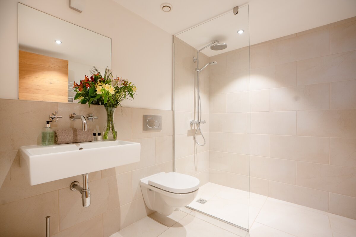 A modern, neutral-colored bathroom with a sleek sink, mirror, decorative flowers, wall-mounted toilet, and a glass walk-in shower.