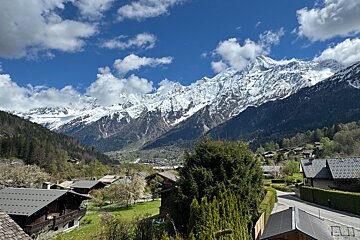 A picturesque village nestled in a green valley, framed by stunning snow-capped mountains under a clear blue sky with fluffy clouds.