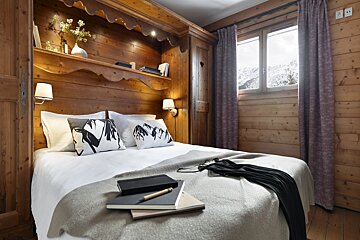 A cozy, rustic wooden bedroom features a bed with mountain-themed pillows, books, and a window revealing a snowy mountain landscape.