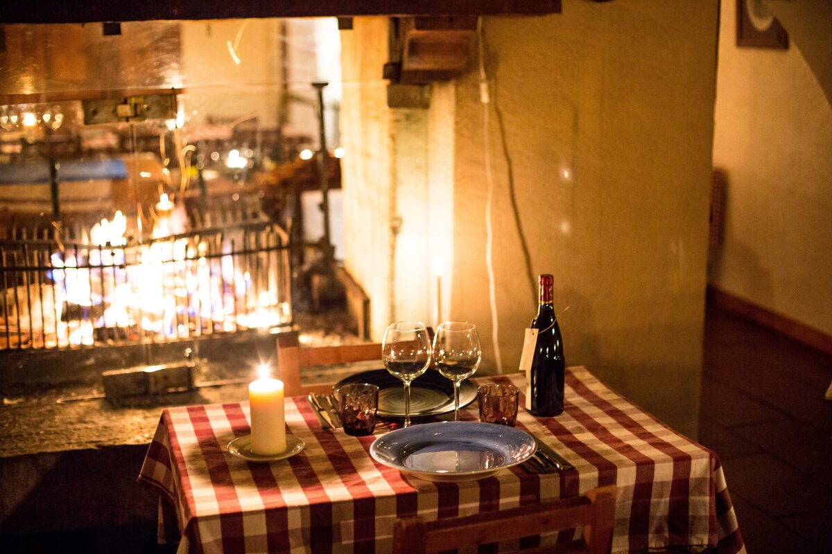 A cozy, candlelit table for two with wine and a checkered tablecloth, set against the warm glow of a roaring fireplace, creating a romantic ambiance.