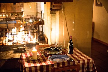 A cozy, candlelit table for two with wine and a checkered tablecloth, set against the warm glow of a roaring fireplace, creating a romantic ambiance.