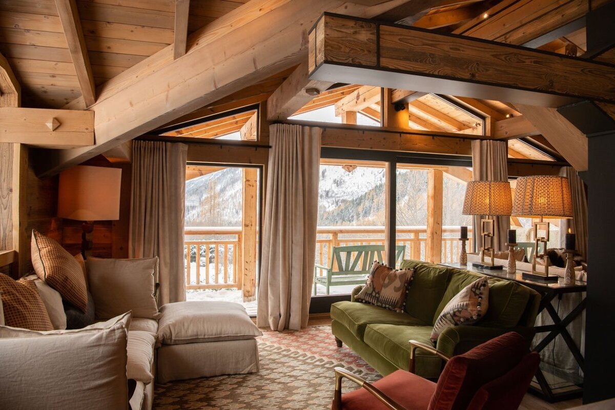 Luxurious wood-paneled chalet living room with cozy sofas, warm lighting, and large windows revealing a snowy mountain vista.