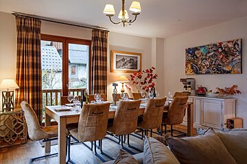 A cozy dining room with a long wooden table set for guests. Brown chairs line the table, while plaid curtains frame a balcony view of a village. Warm lighting and art adorn the space.