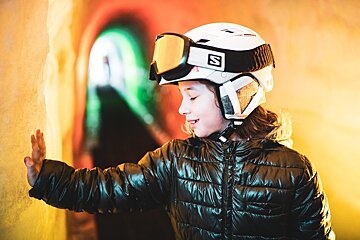 A child in a ski helmet and goggles smiles, hand on a yellow wall, inside a vibrant, glowing tunnel.