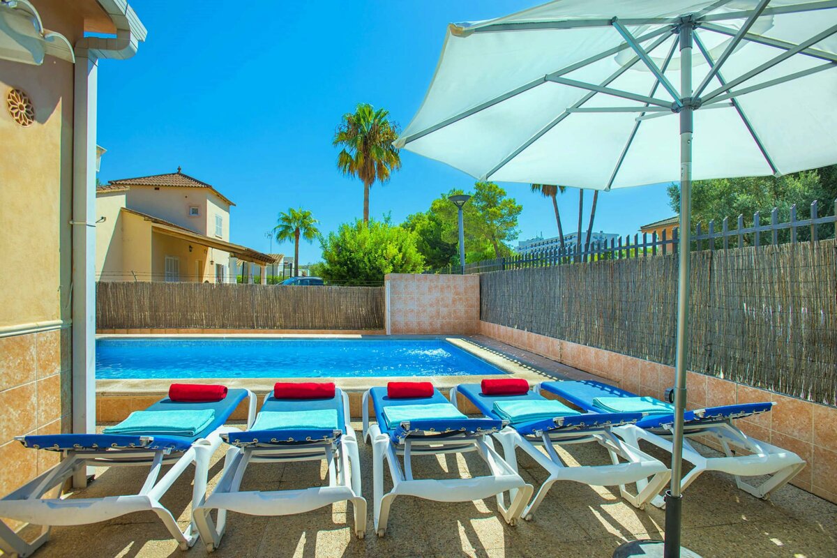 A row of lounge chairs sit under an umbrella in front of a swimming pool