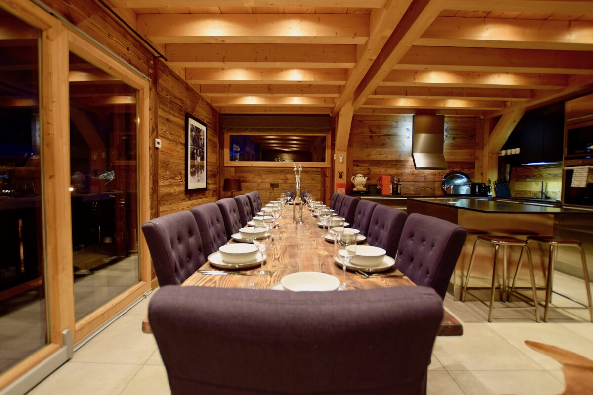 A luxurious chalet-style dining room & kitchen with a long table set for many. Features include wood paneling, exposed beams, and plush purple chairs.
