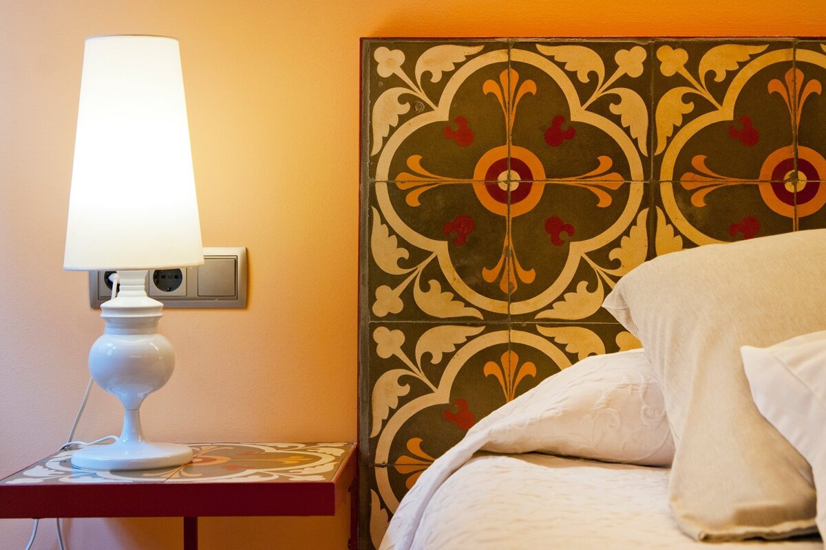 A bedroom scene with a glowing white lamp on a tiled nightstand, next to a bed with cream pillows and an ornate, patterned tiled headboard.