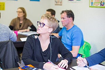 A woman wearing glasses sits at a desk in a classroom