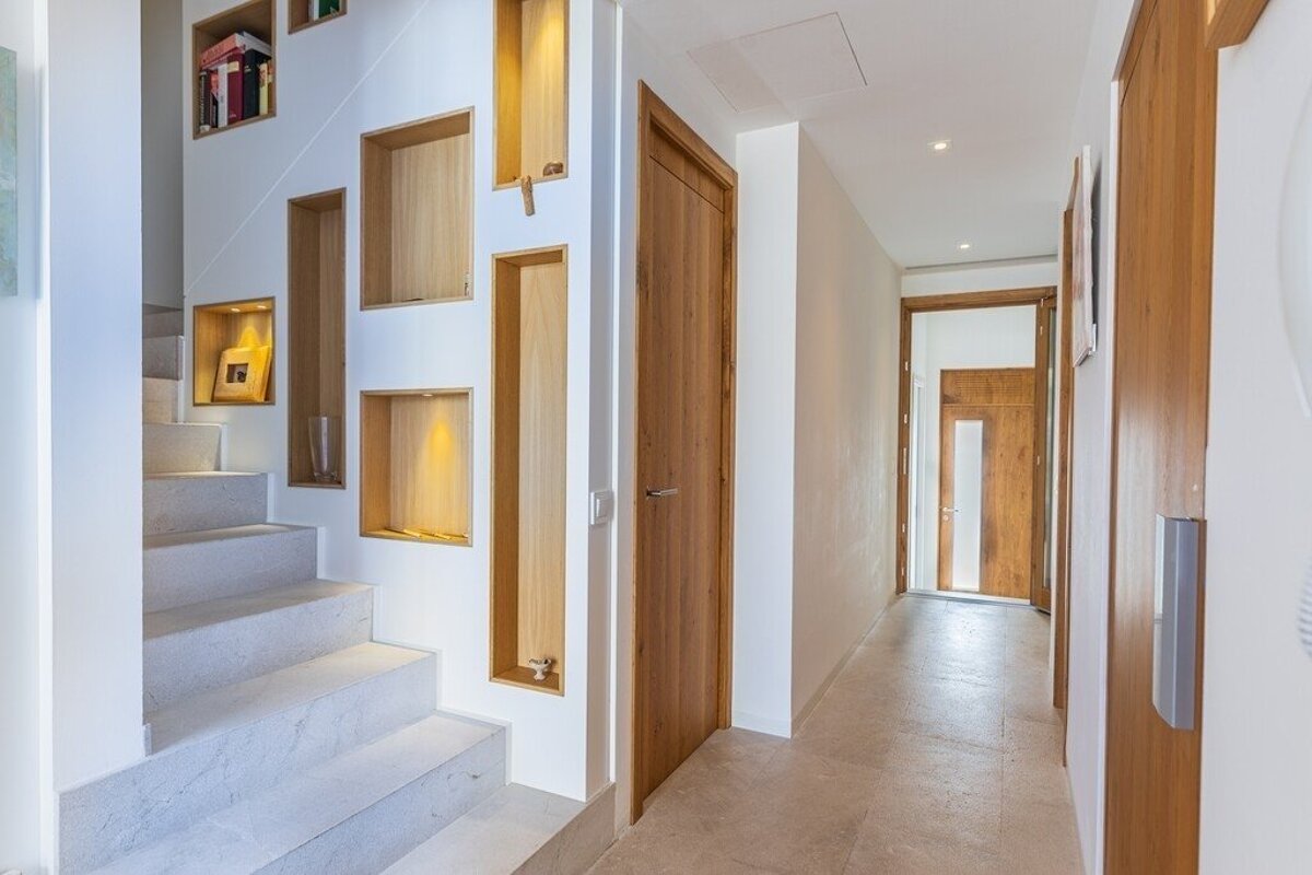 Bright modern interior with a stone staircase, illuminated wall niches, and a hallway with multiple wooden doors.