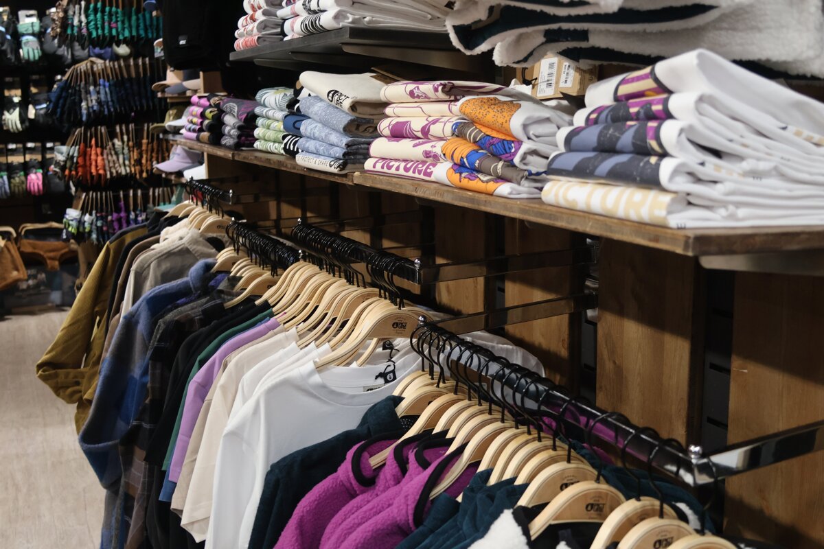A retail store displays racks of hanging clothing, including shirts and jackets, alongside shelves neatly stacked with folded apparel and accessories like gloves.