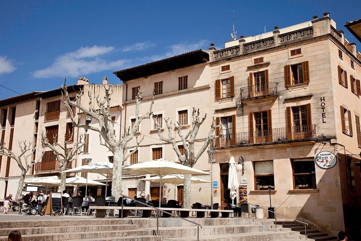 Sunny town square with a hotel, cafe, outdoor seating, bare trees, and historic buildings under a clear blue sky.