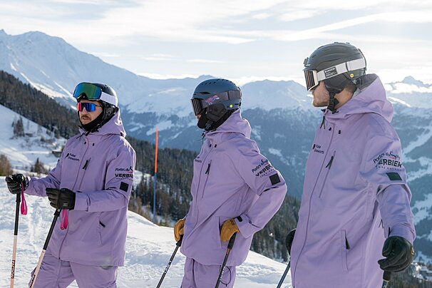 Three skiers in matching purple suits and helmets stand on a sunny, snowy mountain slope, looking out at the stunning view.