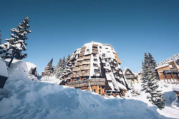 A unique, snow-covered, dome-shaped building with wooden balconies stands among snowy pine trees under a clear blue sky in a winter mountain village.