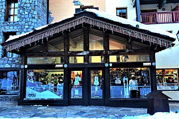 A snow-covered ski rental shop with a wooden facade, large windows, and icicle lights, displaying winter sports gear in a snowy setting.
