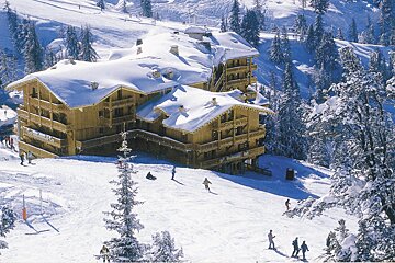 A large, snow-covered wooden ski lodge sits on a snowy mountain slope with skiers and snow-laden pine trees.