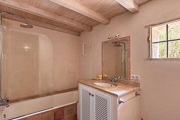A rustic bathroom with a bathtub/shower, vanity, and mirror. It features wooden ceiling beams, a small window, and warm, earthy tones.
