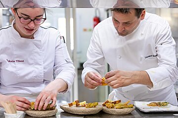 A man and a woman are preparing food in a kitchen and one of the chefs has laurent bord written on his jacket
