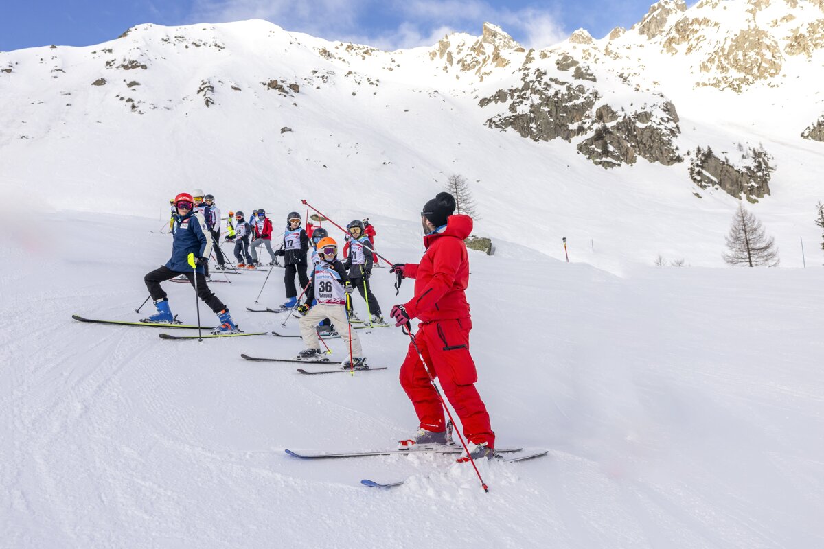 A group of skiers, including children with numbered bibs, line up on a snowy slope. An instructor in red watches them against snow-capped mountains.