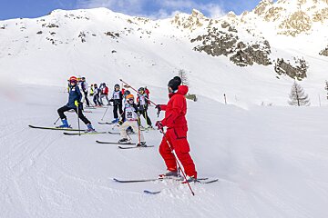 A group of skiers, including children with numbered bibs, line up on a snowy slope. An instructor in red watches them against snow-capped mountains.