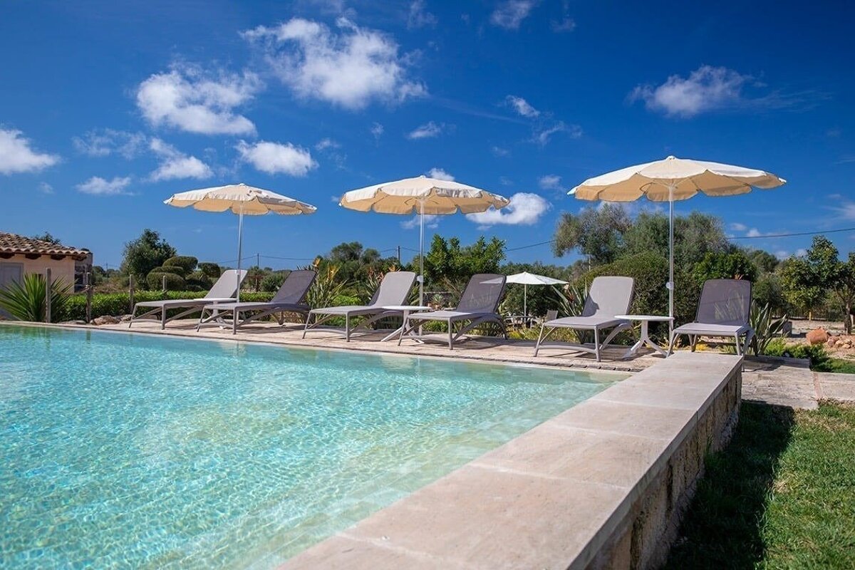 An outdoor swimming pool with several lounge chairs and umbrellas sits under a bright blue sky with white clouds, surrounded by green plants.