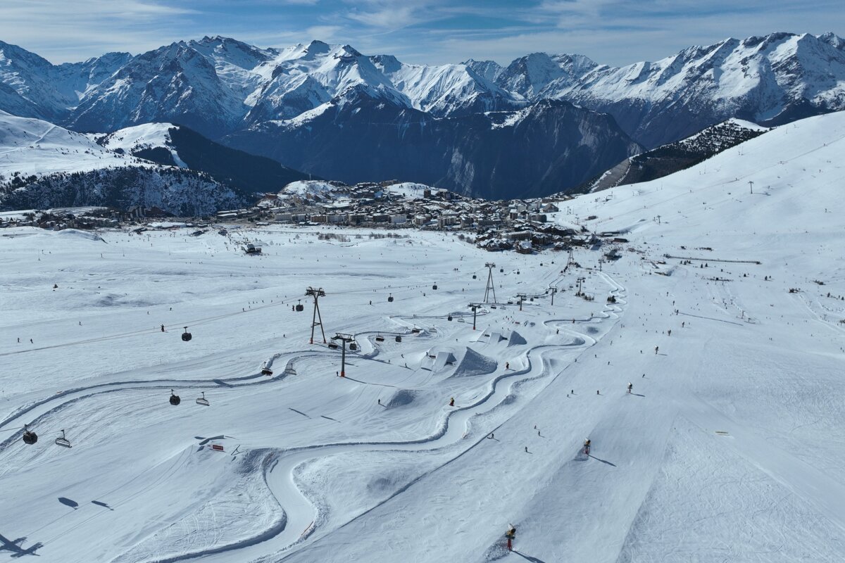 A vibrant ski resort scene with skiers, lifts, and a terrain park in the foreground, with a village and imposing snow-covered mountains beyond.
