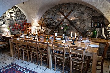 A long wooden dining table set for guests fills a rustic, cellar-like room with stone walls, an arched ceiling, and vintage decor, illuminated by lamps and candles.