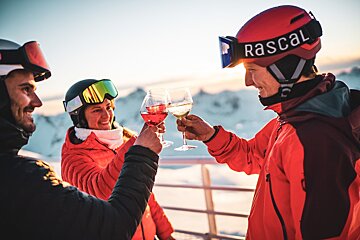 Smiling skiers in helmets and goggles toast with wine on a snowy mountain during golden hour.