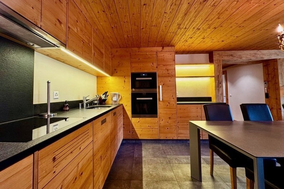 A modern kitchen featuring wooden cabinetry, dark countertops, built-in ovens, and an adjacent dining area.