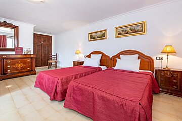 A cozy, traditional bedroom featuring two single beds with red covers, ornate wooden furniture, white walls, and framed art.
