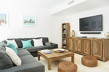 Modern living room with a grey sectional sofa, wooden coffee table, leather poufs, and a TV mounted over a wooden sideboard.