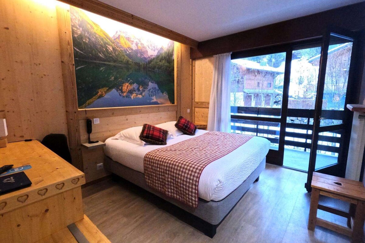 A cozy, wood-paneled chalet-style hotel room features a large bed, a scenic mountain mural, and a balcony view of a snowy landscape.