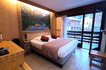 A cozy, wood-paneled chalet-style hotel room features a large bed, a scenic mountain mural, and a balcony view of a snowy landscape.