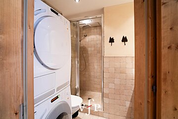 A compact bathroom featuring a stacked washer/dryer unit, a glass shower stall, and a toilet. Tiled walls and wooden accents create a functional space.