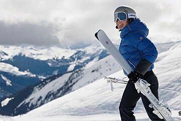 A smiling skier in a blue jacket, helmet, and goggles carries her skis against a snowy mountain backdrop.