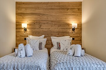 A cozy bedroom with two twin beds, a rustic wooden headboard, warm wall-mounted lamps, and white bathrobes/towels laid out on each bed.