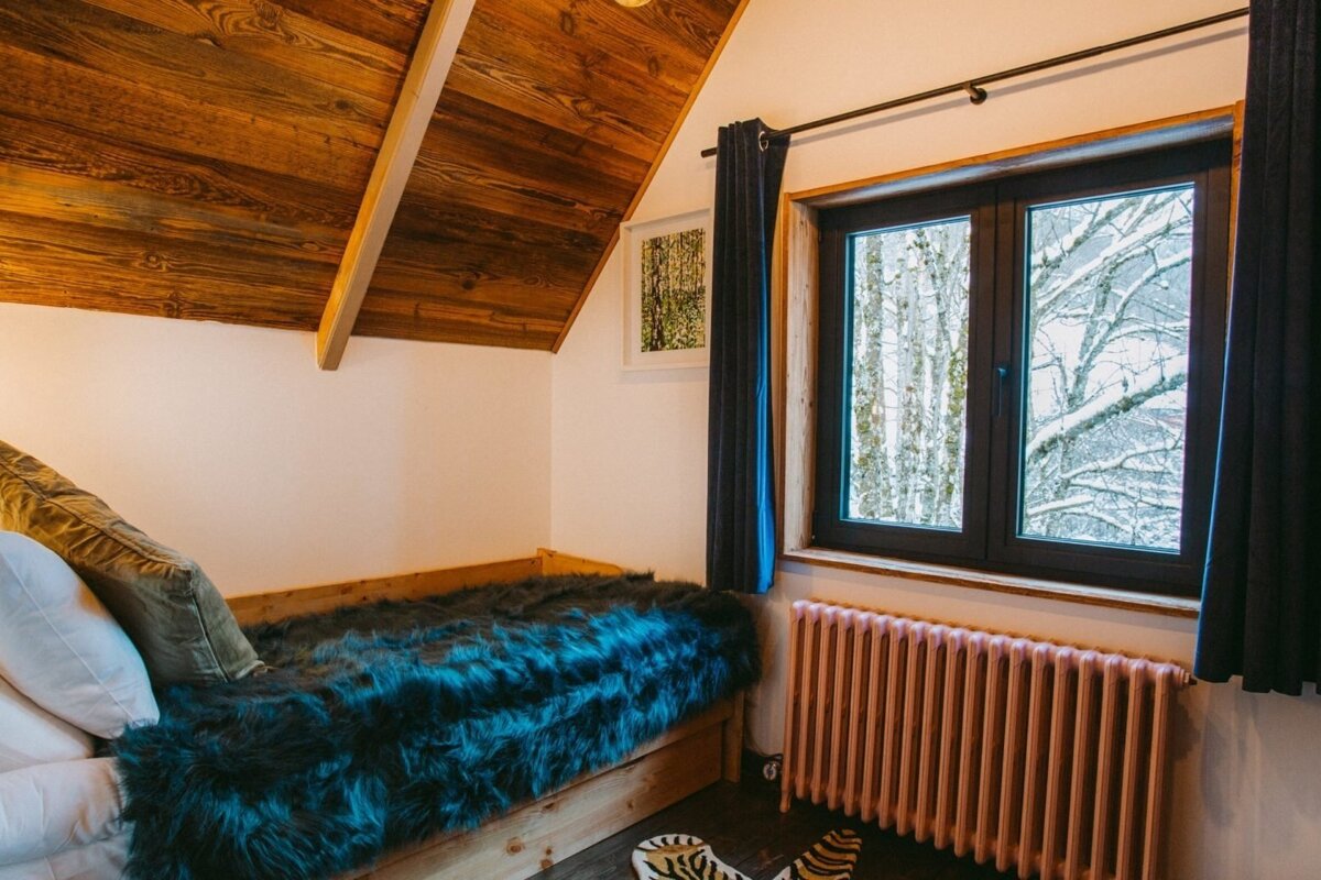 Cozy attic bedroom features a wooden ceiling, bed with faux fur blanket, and a window view of snowy trees above a radiator.