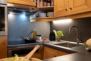 A cozy kitchen counter with wooden cabinets, a sink, an induction stove with a pan, sausages on a plate, and warm under-cabinet lighting.